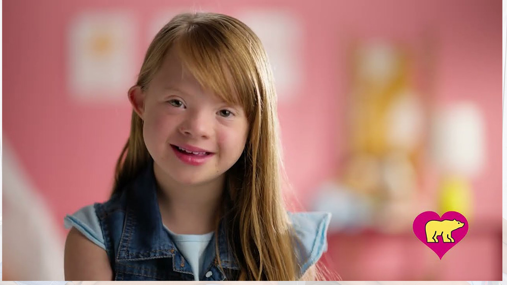 girl with down syndrome smiles in front of pink background. click to watch video.
